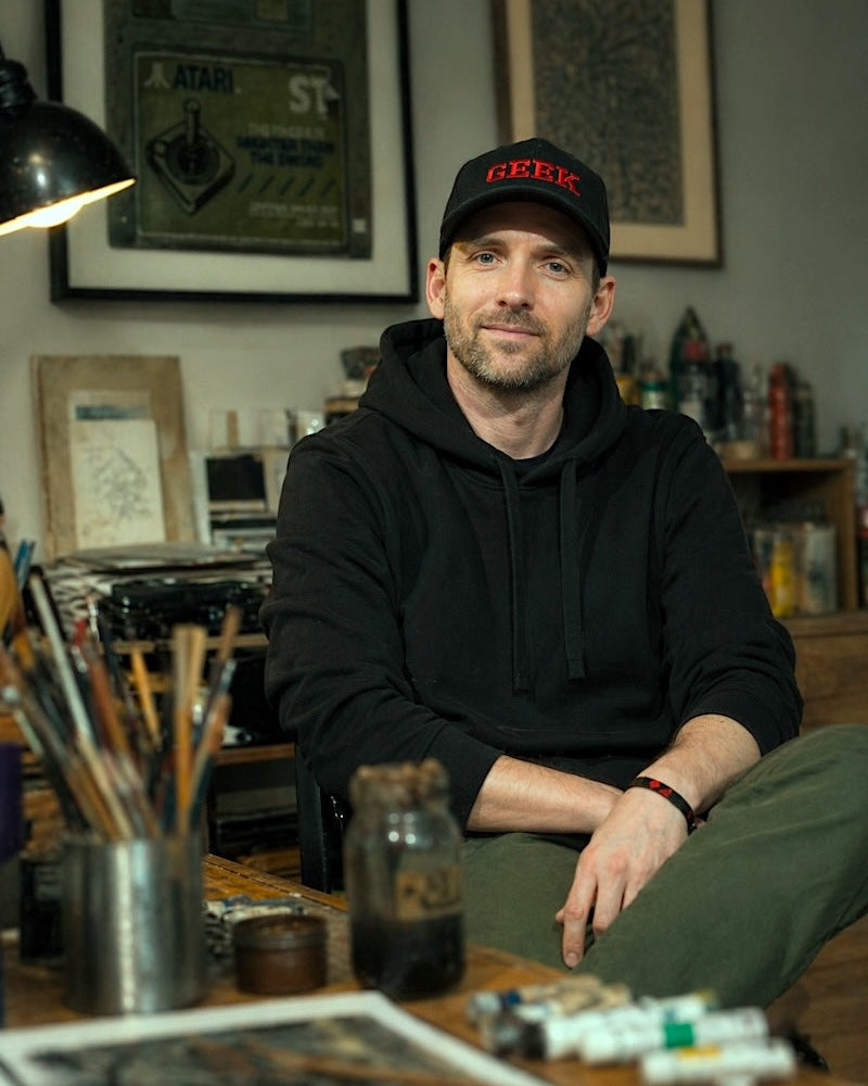 Arlo Sinclair sitting at a desk with art supplies, wearing a black hoodie and cap.